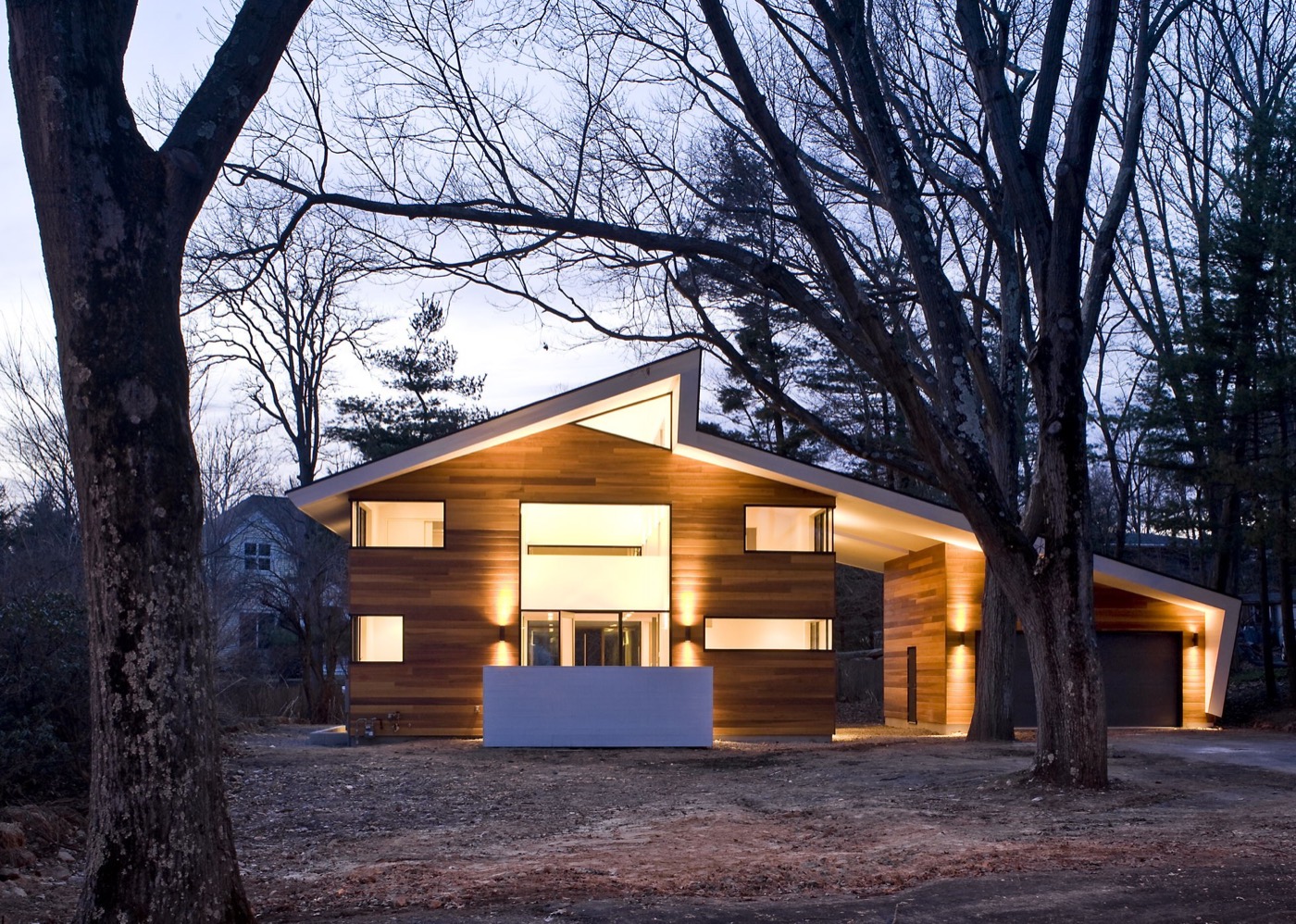 A low modern house seen through bare winter trees at dusk, its interior lit warm, the white underside of a folded roof plane glowing above a white entry wall