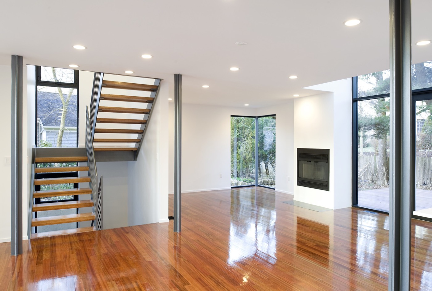Unfurnished ground floor: an open steel-and-wood stair at left, thin steel columns, a fireplace, corner glazing to woods, a glossy cherry floor