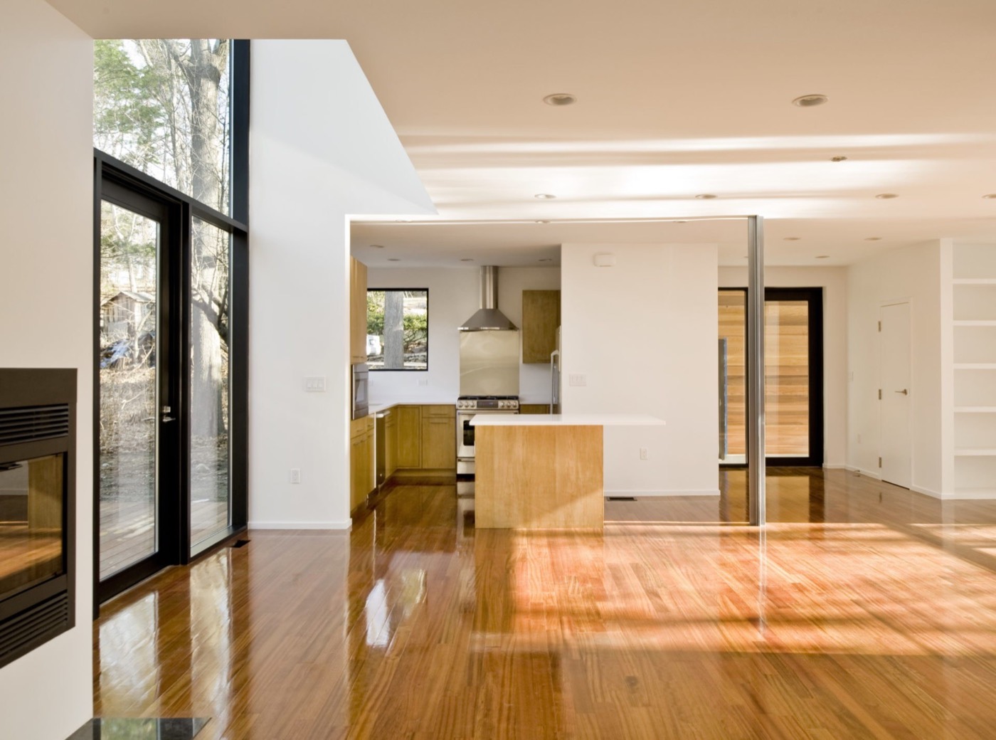 Ground floor looking toward a white kitchen island: fireplace at left, full-height glazing to a deck, steel columns, built-in shelving