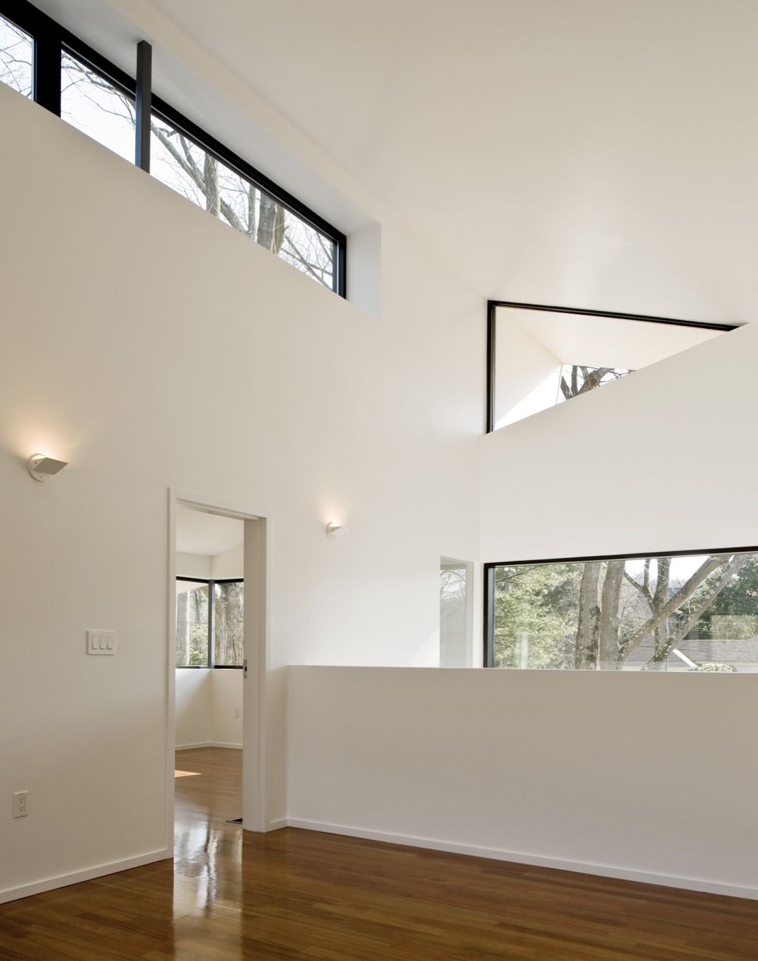 Upper level of the double-height space: white walls, a clerestory ribbon window, black-framed openings, a low guard wall, a bedroom door beyond