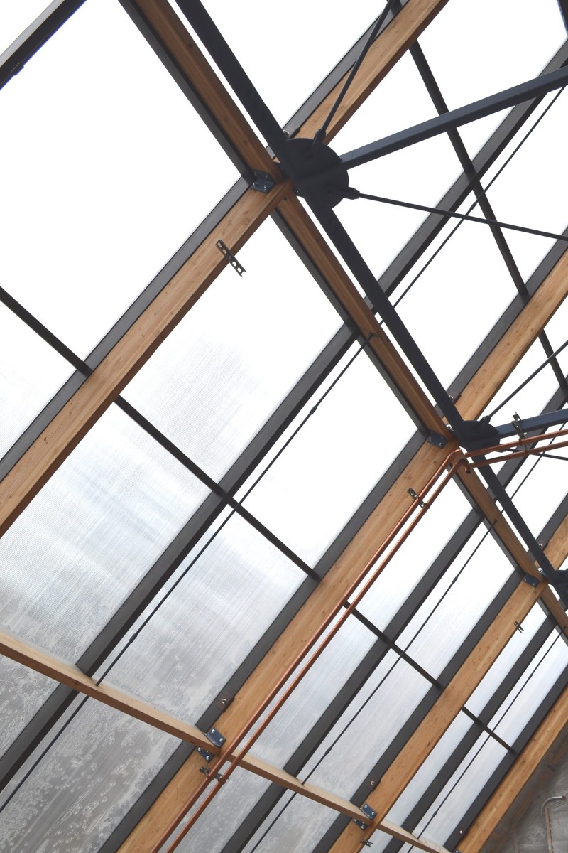 Interior view up through the sloped glazing frame: wood rafters, steel tension rods meeting a cast node, translucent panels, a copper pipe run