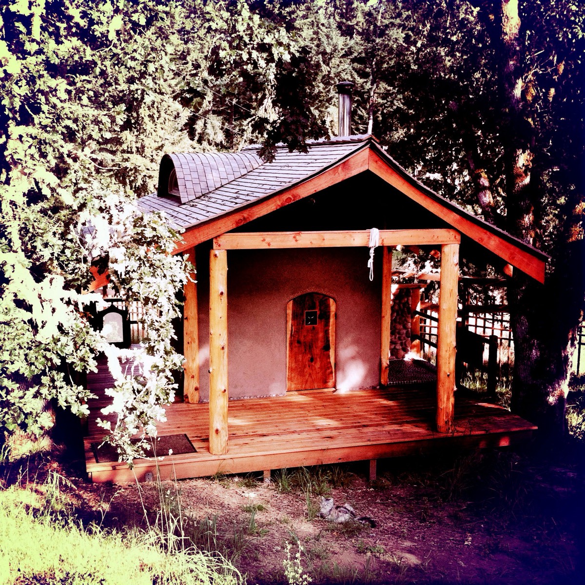 The front of the sauna: an arched hand-made timber door centred in an earthen wall, four log posts, the shingled roof and skylight above, a deck below