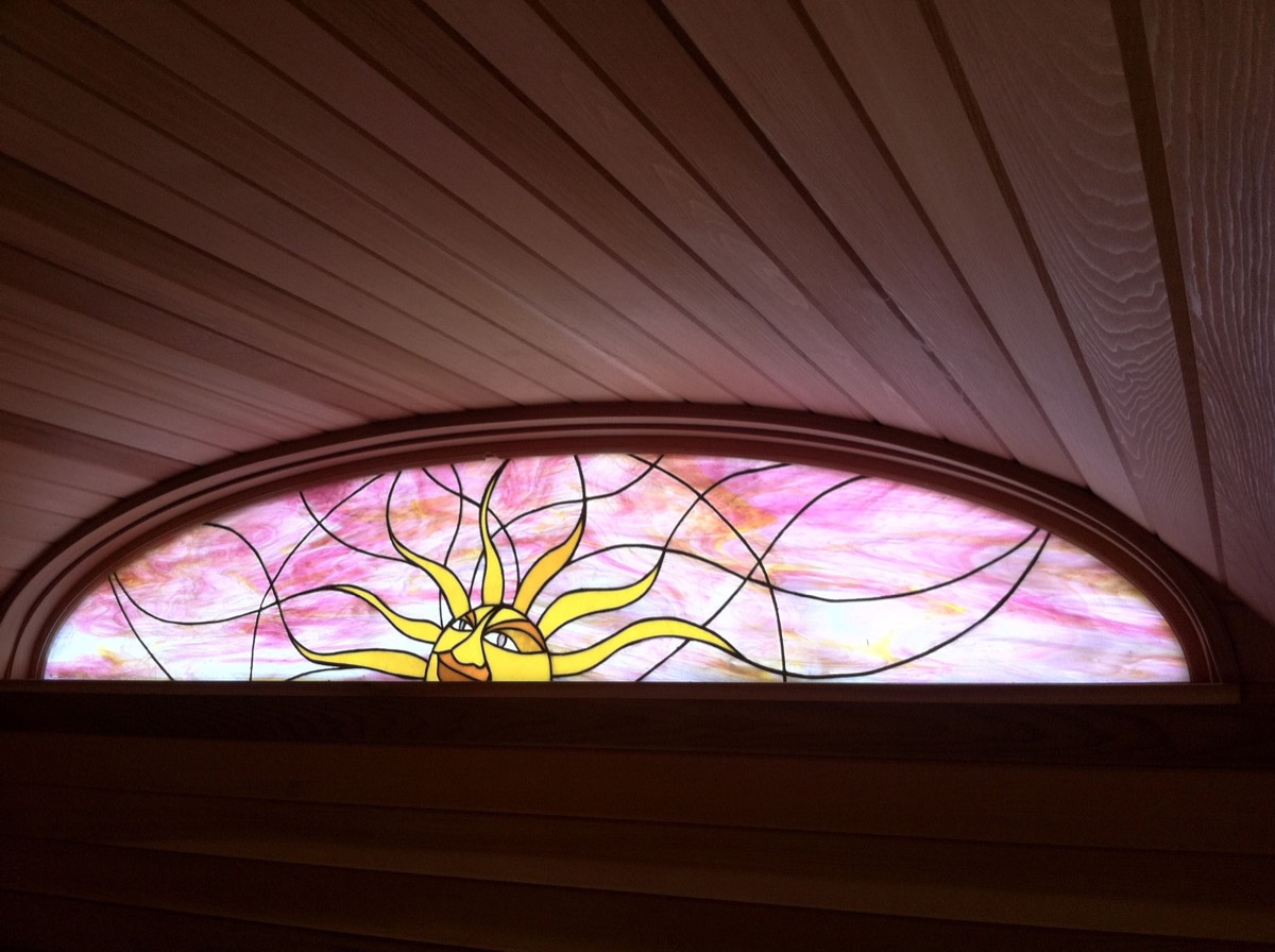 Looking up at an arched stained-glass window set into a curved cedar ceiling: a yellow sun face on a pink marbled ground