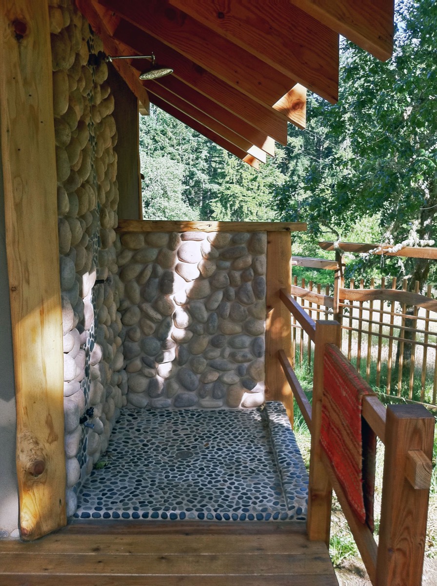 An outdoor shower of river-cobble walls and a black-pebble mosaic floor, a brass shower head under exposed rafter tails, forest beyond