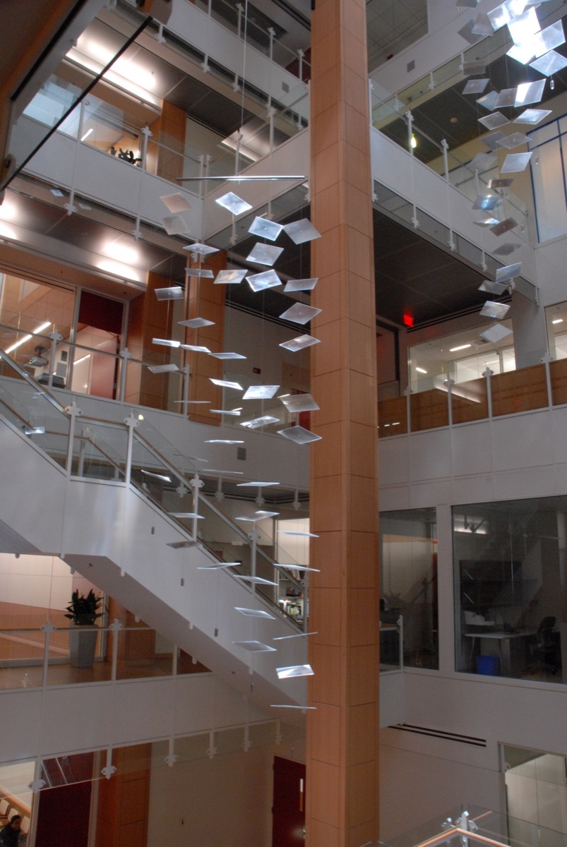 A tall view of one mobile of clear plates spiralling down an eight-storey atrium beside a stair and a wood-clad column