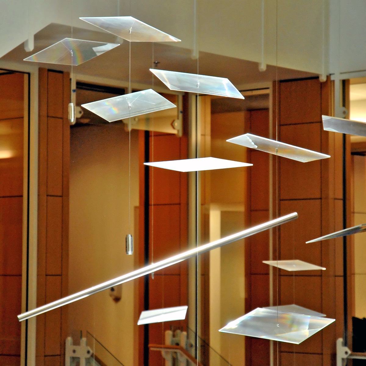 A dozen acrylic plates on monofilament with small counterweights, rainbow spectra across their faces, a warm wood wall behind
