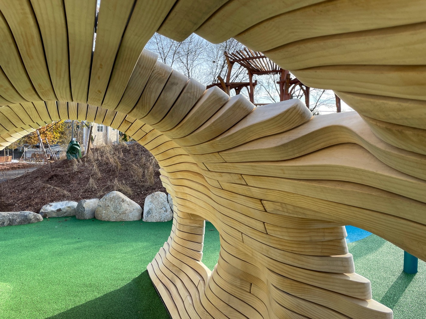 From inside the pipe looking out through its broken end: wave-cut plank strata sweeping across the frame, a pergola and bare trees beyond