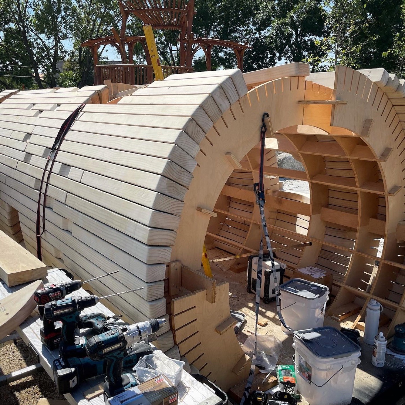 A plywood rib jig standing open in the park with shaped planks stacked around it, drills and buckets on a bench in front