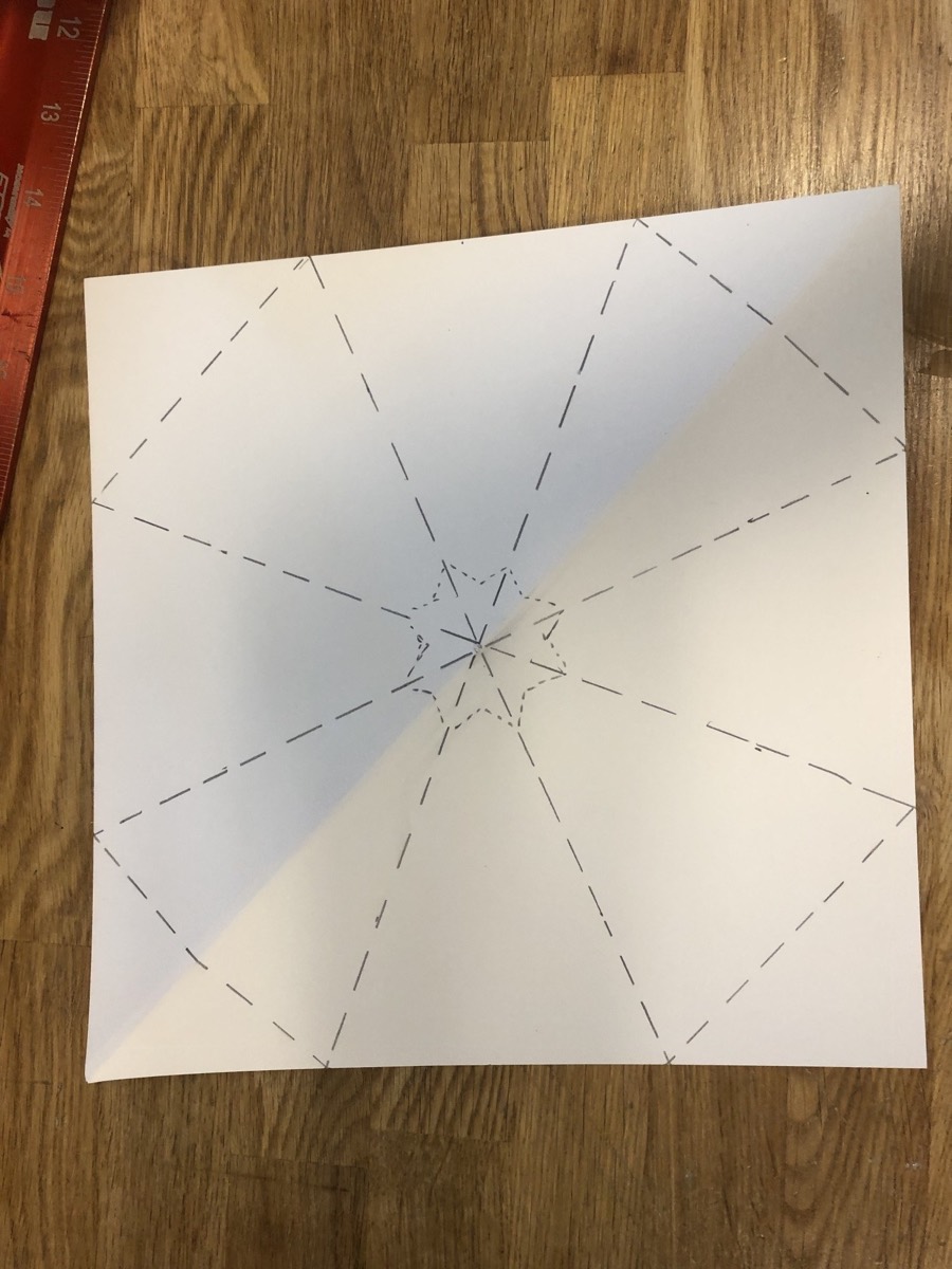 A square of paper scored with radiating dashed lines on a wood floor